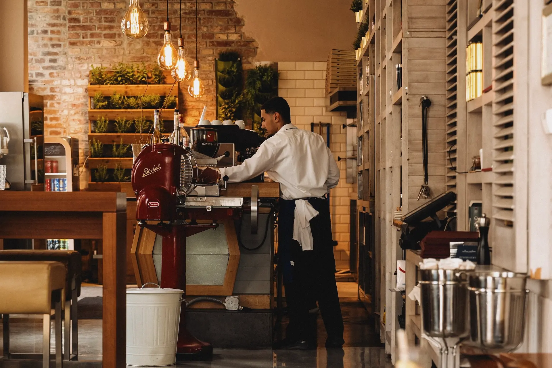 Ein Barista in weißem Hemd und Schürze bereitet an einer Theke neben einer roten Aufschnittmaschine Kaffee zu.