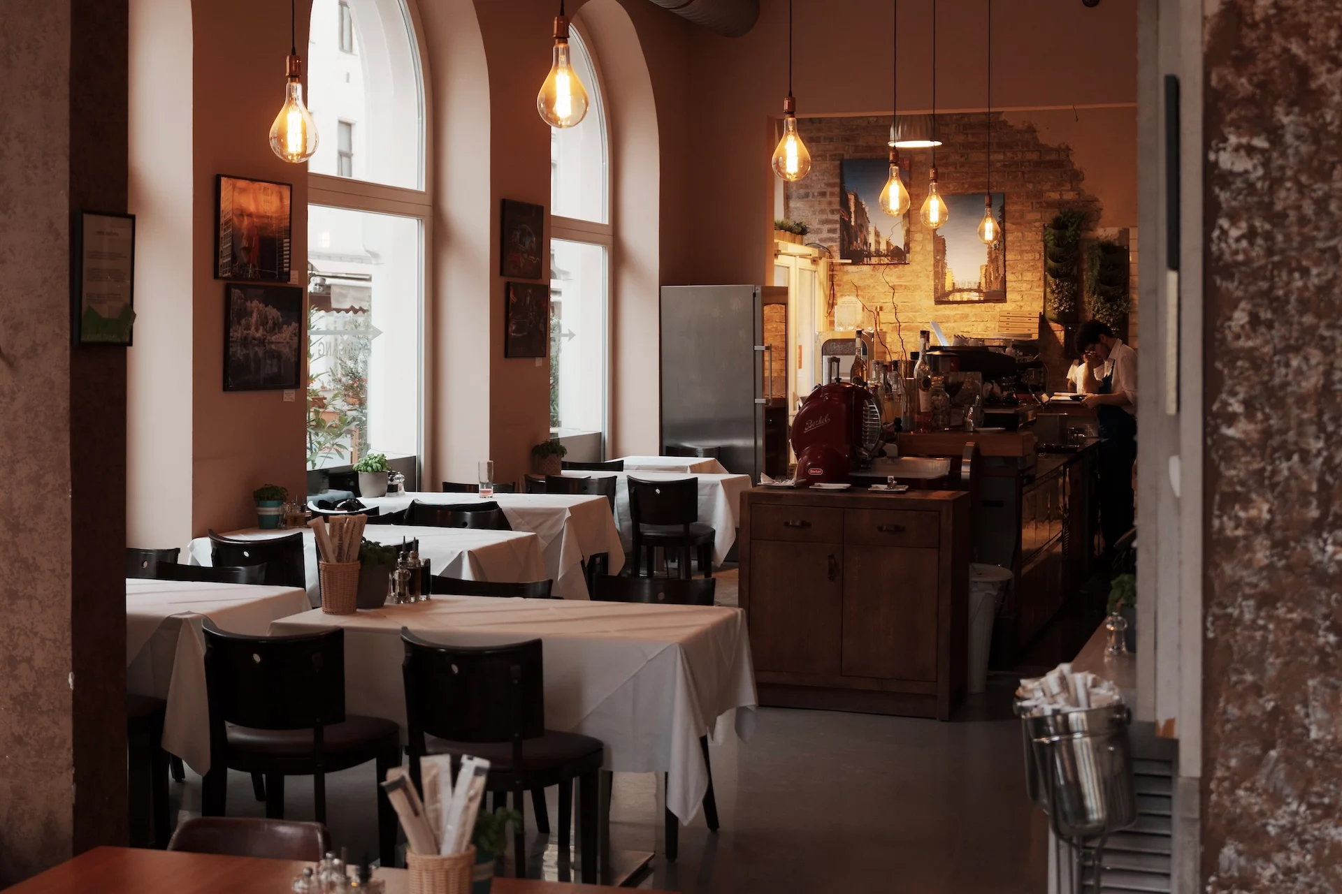 Tische mit weißen Tischdecken und dunklen Stühlen in einem schwach beleuchteten Café mit Hängeleuchten.
