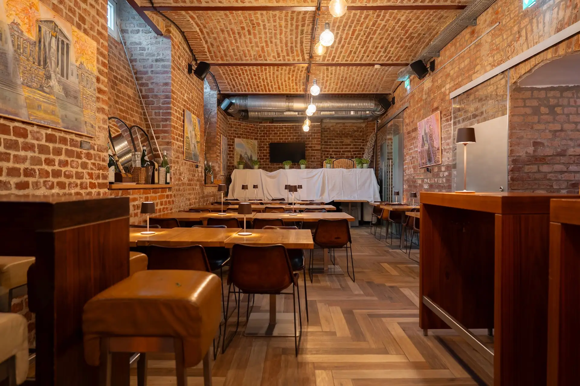 Innenraum eines Restaurants mit Backsteinwänden, Reihen von Holztischen mit kleinen Lampen und Fischgrätenparkett.