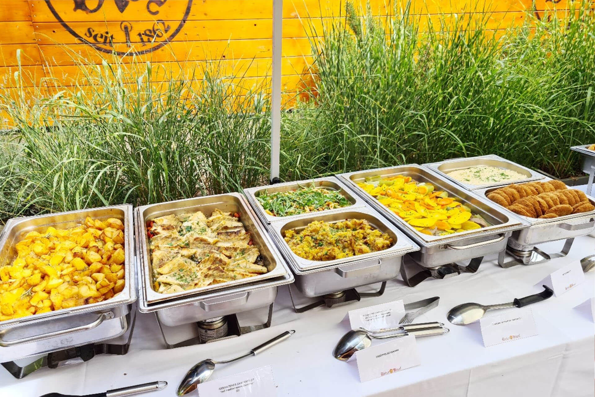 Auf einem Büffettisch im Freien stehen Tabletts mit warmen Speisen, darunter Bratkartoffeln, Nudeln, Gemüse, in Scheiben geschnittener gelber Kürbis und eine Cremeschale mit Keksen, alles fein säuberlich mit Servierutensilien und Etiketten angeordnet.