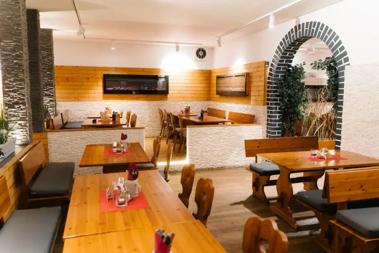 Ein gemütliches, modernes Restaurant mit Holztischen und -bänken, weißen Steinwänden, dekorativen Pflanzen und einem gemauerten Bogengang. Die Tische sind mit roten Tischsets und Geschirr gedeckt, und der Raum ist sanft beleuchtet.