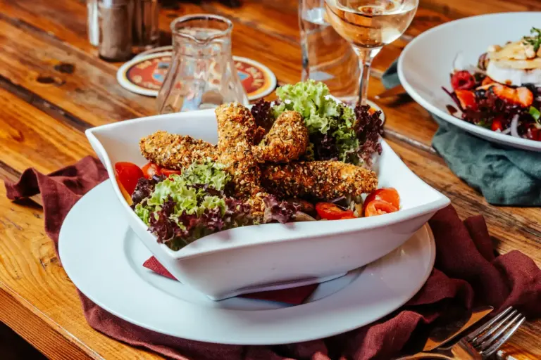Eine Schüssel mit frischem Salat, panierten, gebratenen Käsestangen, Blattgemüse und Kirschtomaten steht auf einem Teller. Auf einem Holztisch im Hintergrund stehen ein Glas Weißwein und ein weiteres Gericht auf einem Teller.