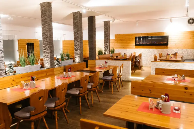 Ein gemütliches Restaurant mit Holztischen und -stühlen, dekorativen Steinsäulen, Pflanzgefäßen und einem modernen elektrischen Kamin an der Wand. Die Tische sind mit Gedecken und roten Tischsets dekoriert.