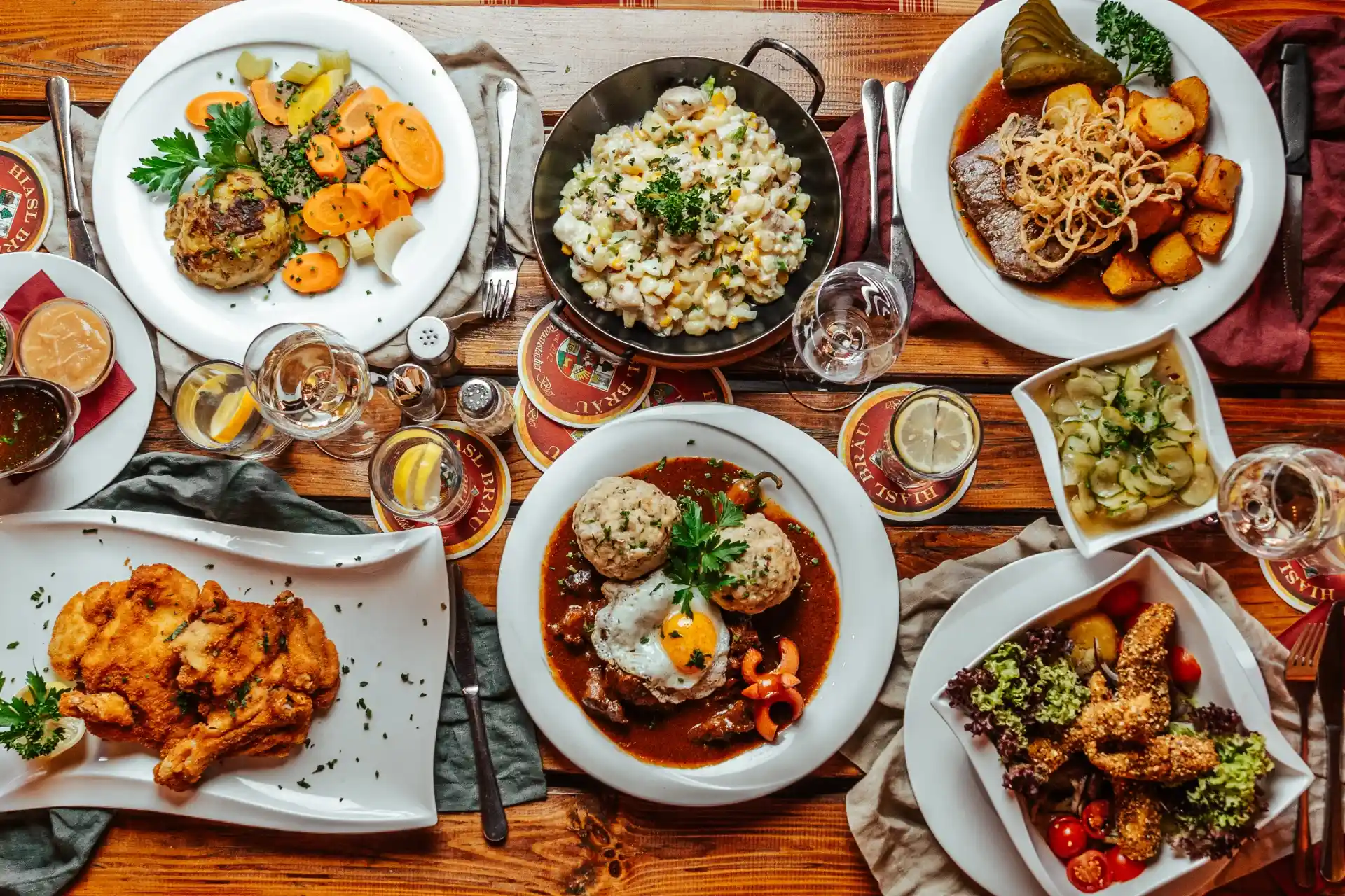 Ein Holztisch mit verschiedenen deftigen deutschen Gerichten, darunter Schnitzel, Kartoffelsalat, Braten mit Soße, Knödel, Gemüse und Gläser mit Wasser und Zitronenscheiben.
