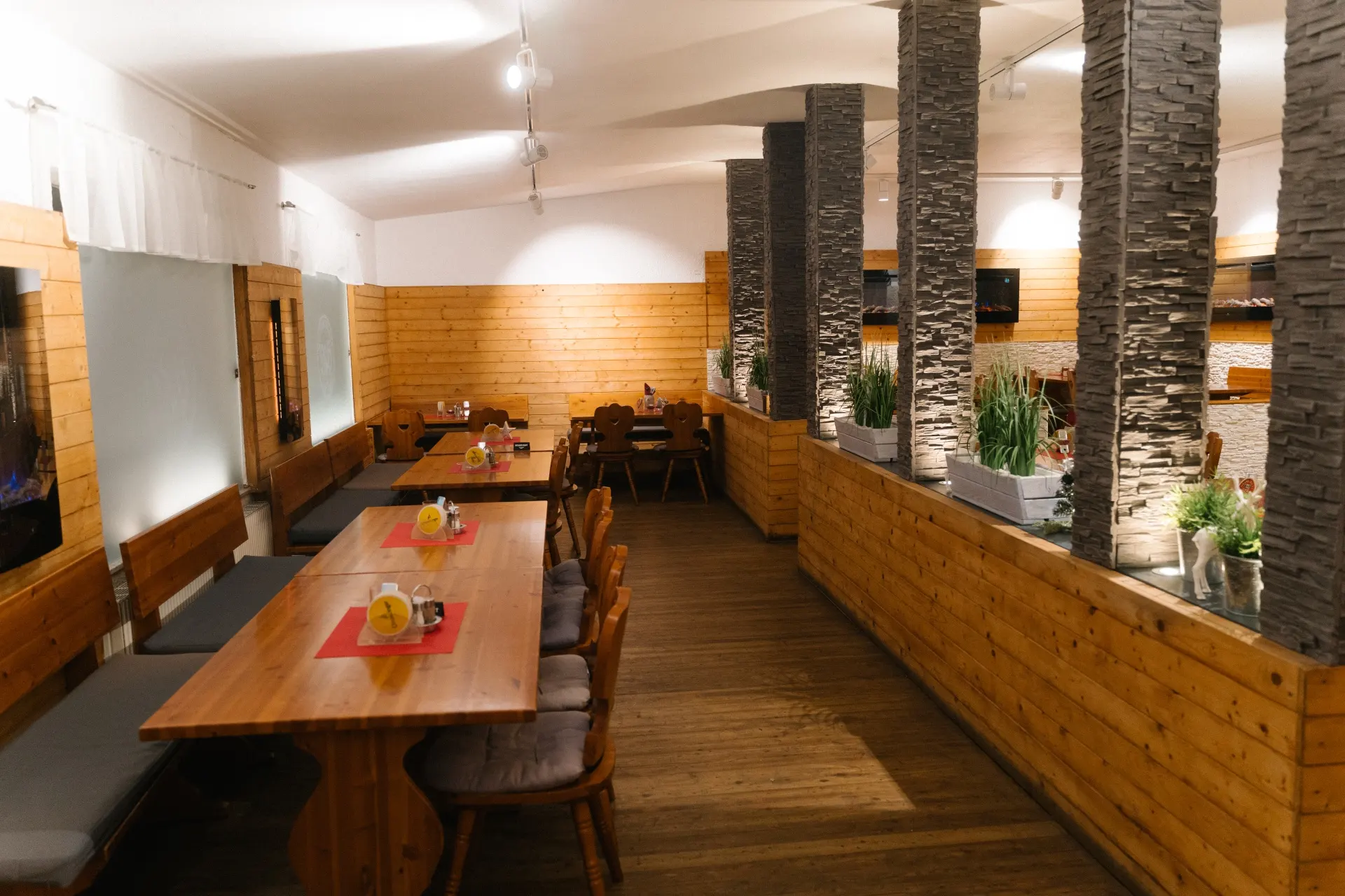 Ein modernes Restaurant mit Holztischen, gepolsterten Stühlen und Steinsäulen als Trennwände. Pflanzen säumen die Trennwand, und eine sanfte Beleuchtung schafft eine gemütliche, einladende Atmosphäre.