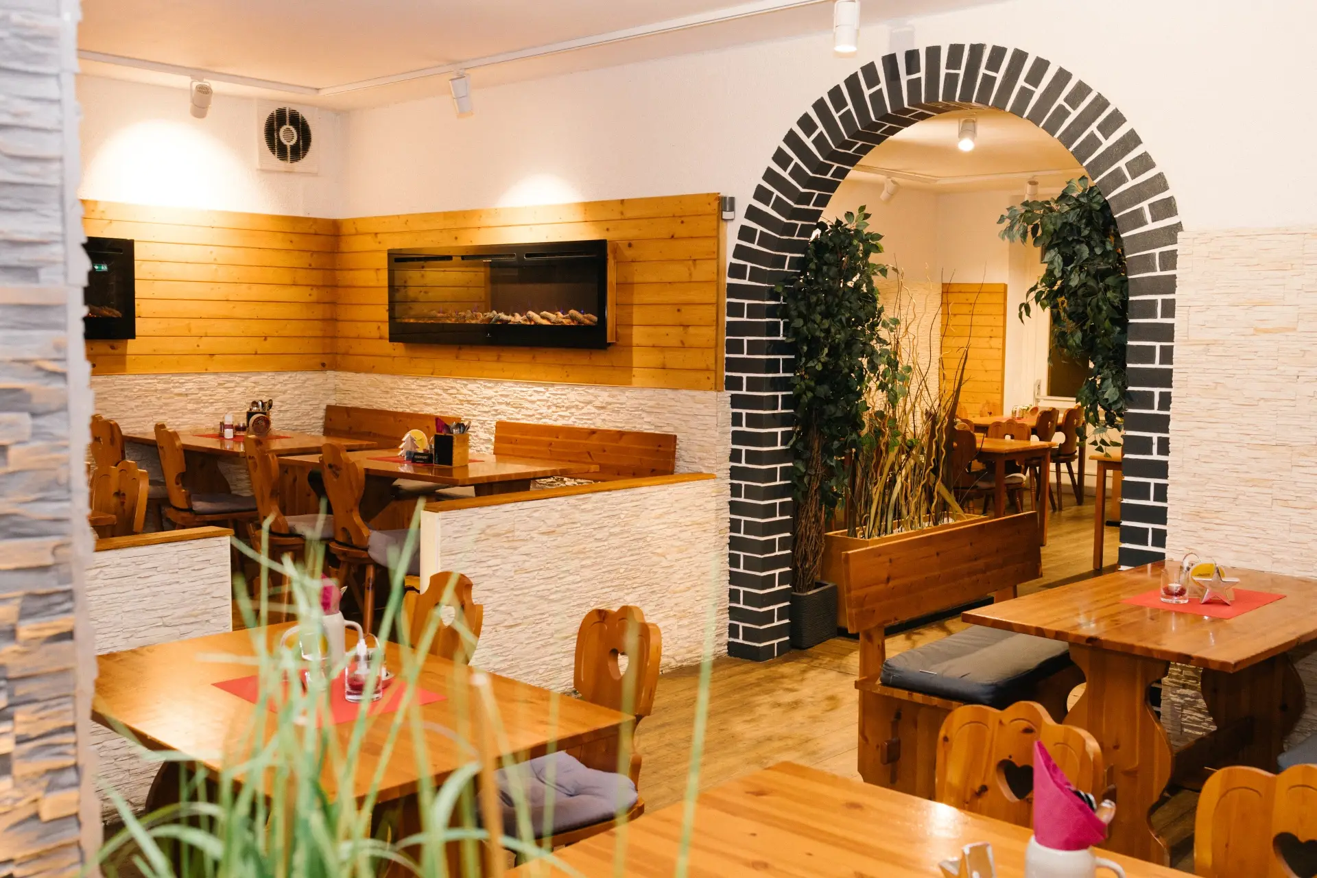 Gemütliches Restaurantinterieur mit Holztischen und -bänken, hellen Wänden und einem schwarz-weißen Backsteinbogen. Grünpflanzen schmücken den Raum und schaffen eine warme und einladende Atmosphäre.