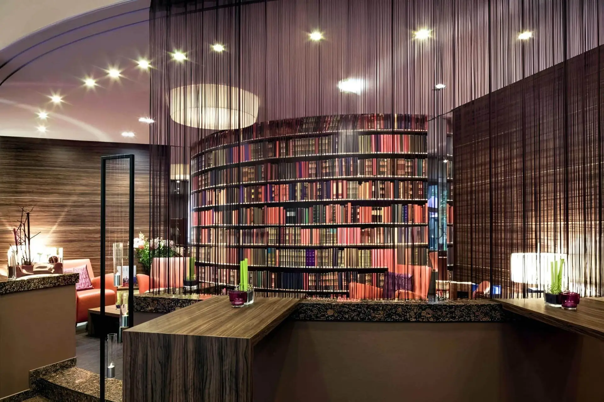 Ein moderner Lounge-Bereich mit einem geschwungenen Bücherregal, das mit bunten Büchern gefüllt ist, schwarzen Vorhängen, die von der Decke baumeln, sanfter Beleuchtung und Holzmöbeln, die eine gemütliche und anspruchsvolle Atmosphäre schaffen.