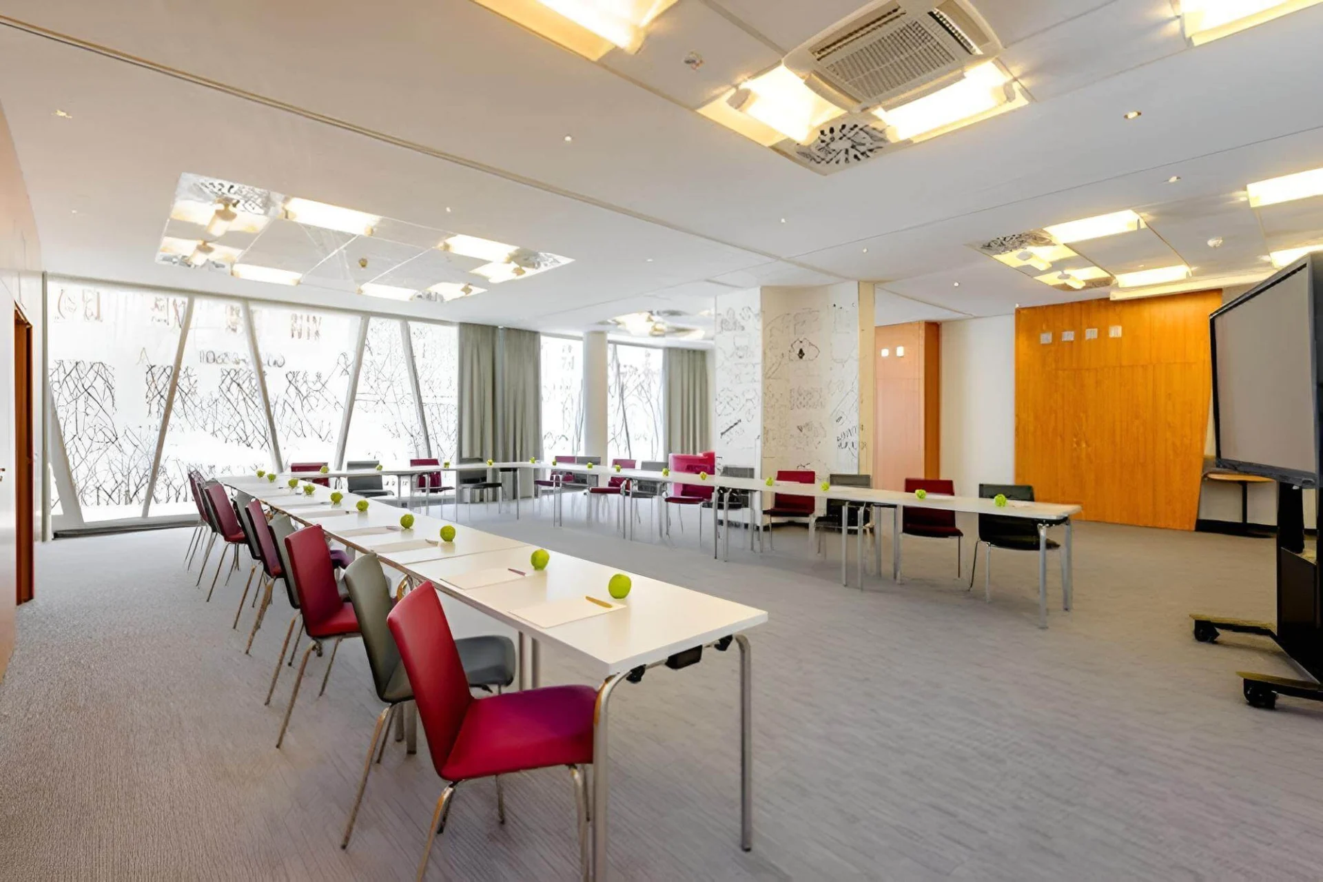 Ein moderner Konferenzraum mit U-förmig angeordneten Tischen, an denen jeweils ein roter Stuhl und ein grüner Apfel auf dem Tisch stehen. Große Fenster lassen natürliches Licht herein, und in einer Ecke steht eine Projektionsfläche.