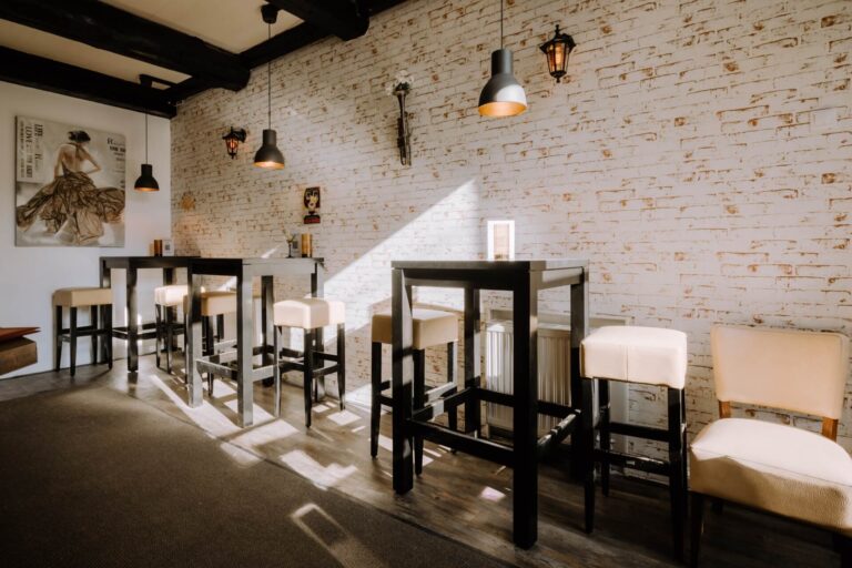 Ein gemütliches Café mit hohen Tischen und cremefarbenen Hockern vor einer weißen Backsteinwand, dekoriert mit modernen Hängelampen und gerahmter Kunst, die von einem nahen Fenster beleuchtet wird.