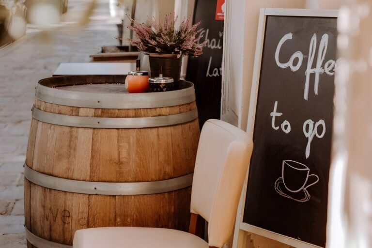 Ein gemütliches Straßencafé mit einem Holzfass als Tisch, einer Topfpflanze und einem Kreideschild mit der Aufschrift "Coffee to go" und einer Tassenillustration, das an einer Kopfsteinpflasterstraße steht.