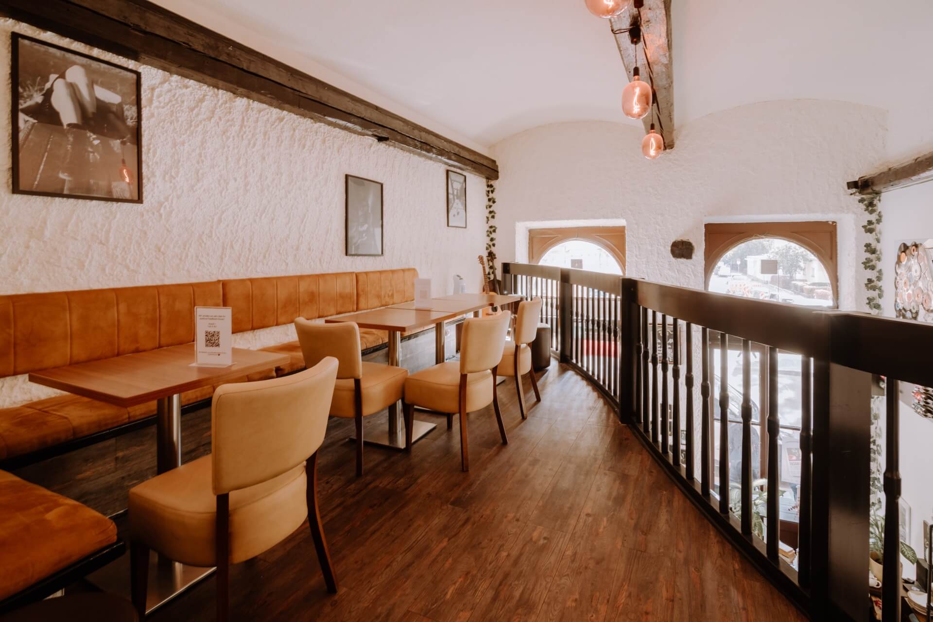 Ein gemütliches Café mit braunen gepolsterten Sitzen, Holztischen und -stühlen. Der Raum hat weiße, strukturierte Wände, gerahmte Kunst, Holzböden und große Bogenfenster, die natürliches Licht hereinlassen.