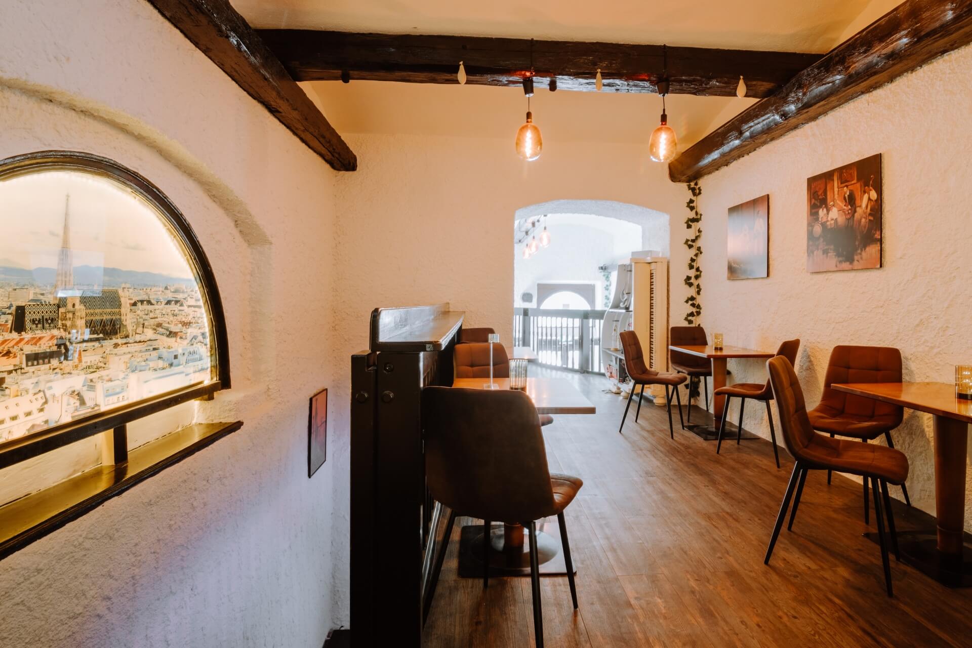 Ein gemütliches Café mit Holzböden, braunen Stühlen und Tischen, freiliegenden Holzbalken und Hängelampen. Auf der linken Seite befindet sich ein Fenster mit Blick auf die Stadt, und an den weißen, strukturierten Wänden hängen gerahmte Kunstwerke.