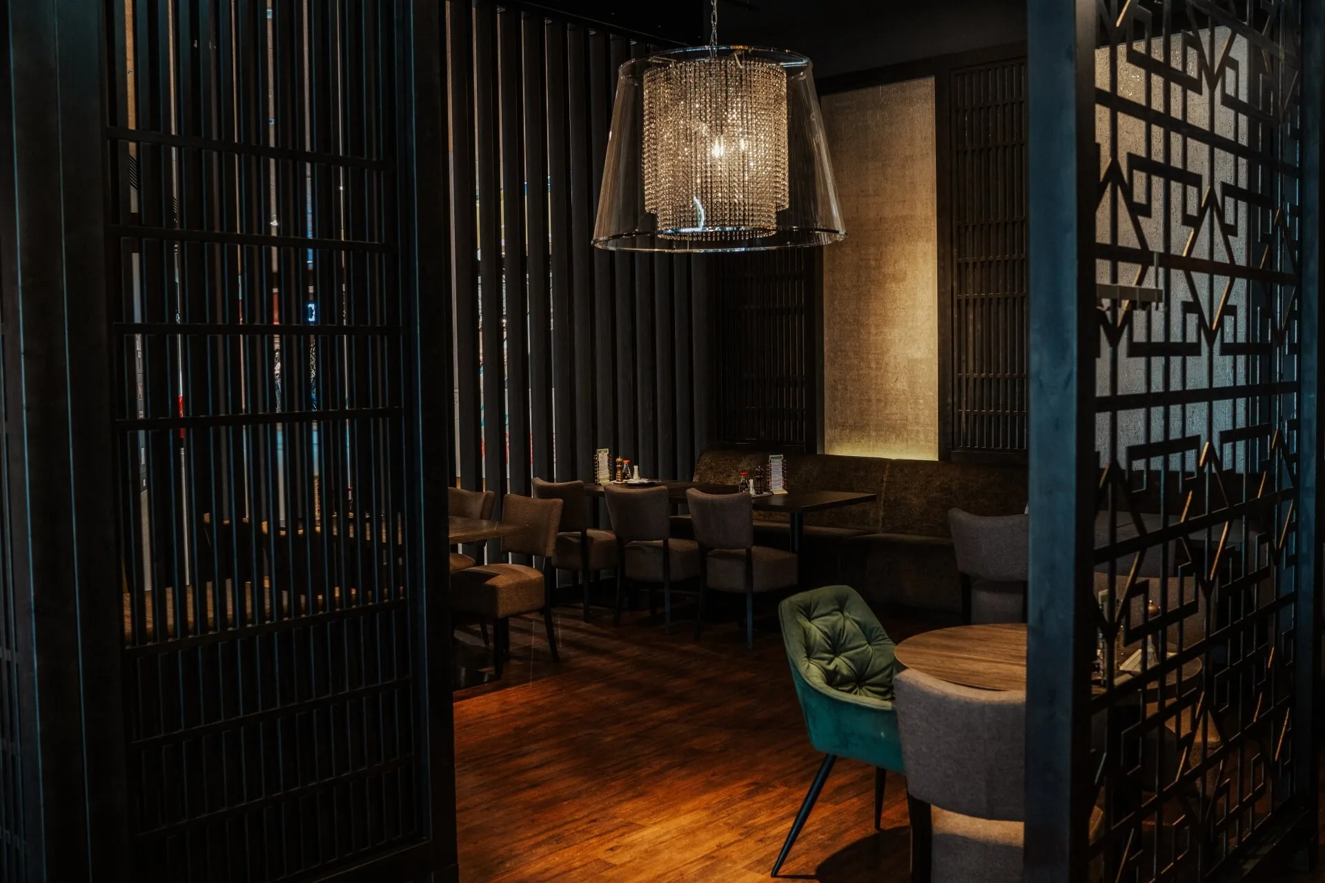 Ein modernes, schummriges Restaurant mit Holzboden, grauen und grünen Stühlen, gedeckten Tischen und einem großen, stilvollen Kronleuchter an der Decke. Der Raum wirkt gemütlich und intim.