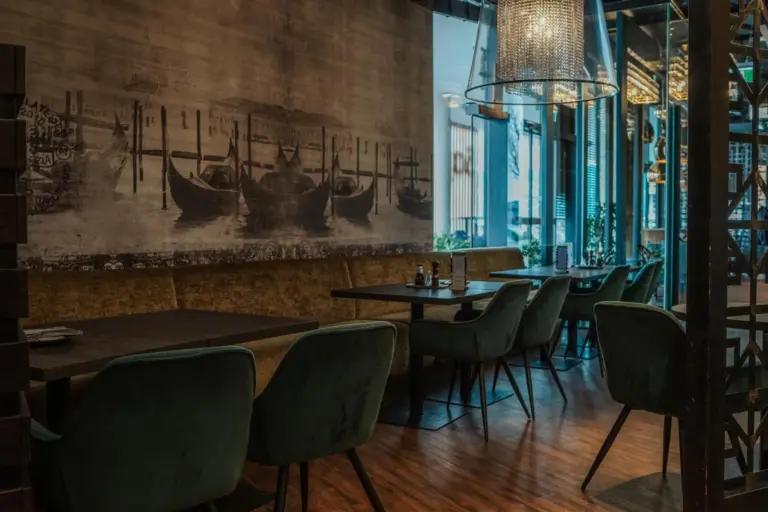 Ein modernes Restaurant mit grünen Samtstühlen, dunklen Holztischen, gedämpfter Beleuchtung und einem großen Wandgemälde mit Gondeln auf dem Wasser, das eine gemütliche und elegante Atmosphäre schafft.
