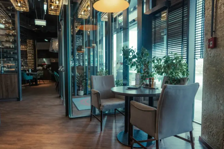 Ein gemütliches Restaurant mit einem runden Tisch und zwei grauen Polsterstühlen in der Nähe großer Fenster, Grünpflanzen, Holzfußboden und moderner Beleuchtung. Der Raum wirkt einladend und ist durch natürliches Licht gut beleuchtet.