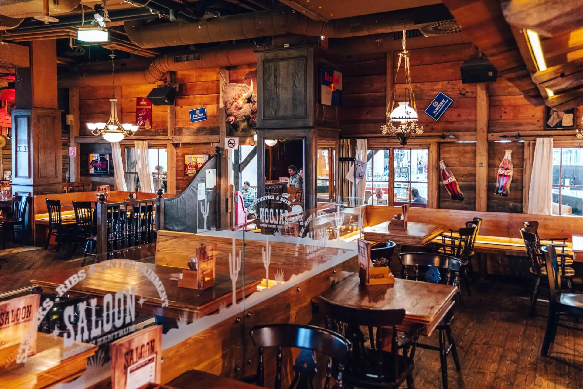 Interieur eines rustikalen Restaurants im Western-Stil mit Holztischen und -stühlen, Vintage-Dekor, Hängelampen, Wandschildern und großen Fenstern, die natürliches Licht hereinlassen. Der Raum wirkt gemütlich und einladend, mit einer entspannten Atmosphäre.