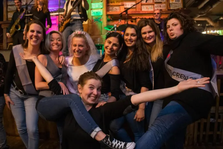 Eine Gruppe lächelnder Frauen posiert zusammen in einem Innenraum auf einer lebhaften Party, wobei eine Frau ein Diadem trägt. Eine Frau streckt spielerisch ihre Arme und Beine nach vorne, während im Hintergrund eine Band unter bunten Lichtern auftritt.