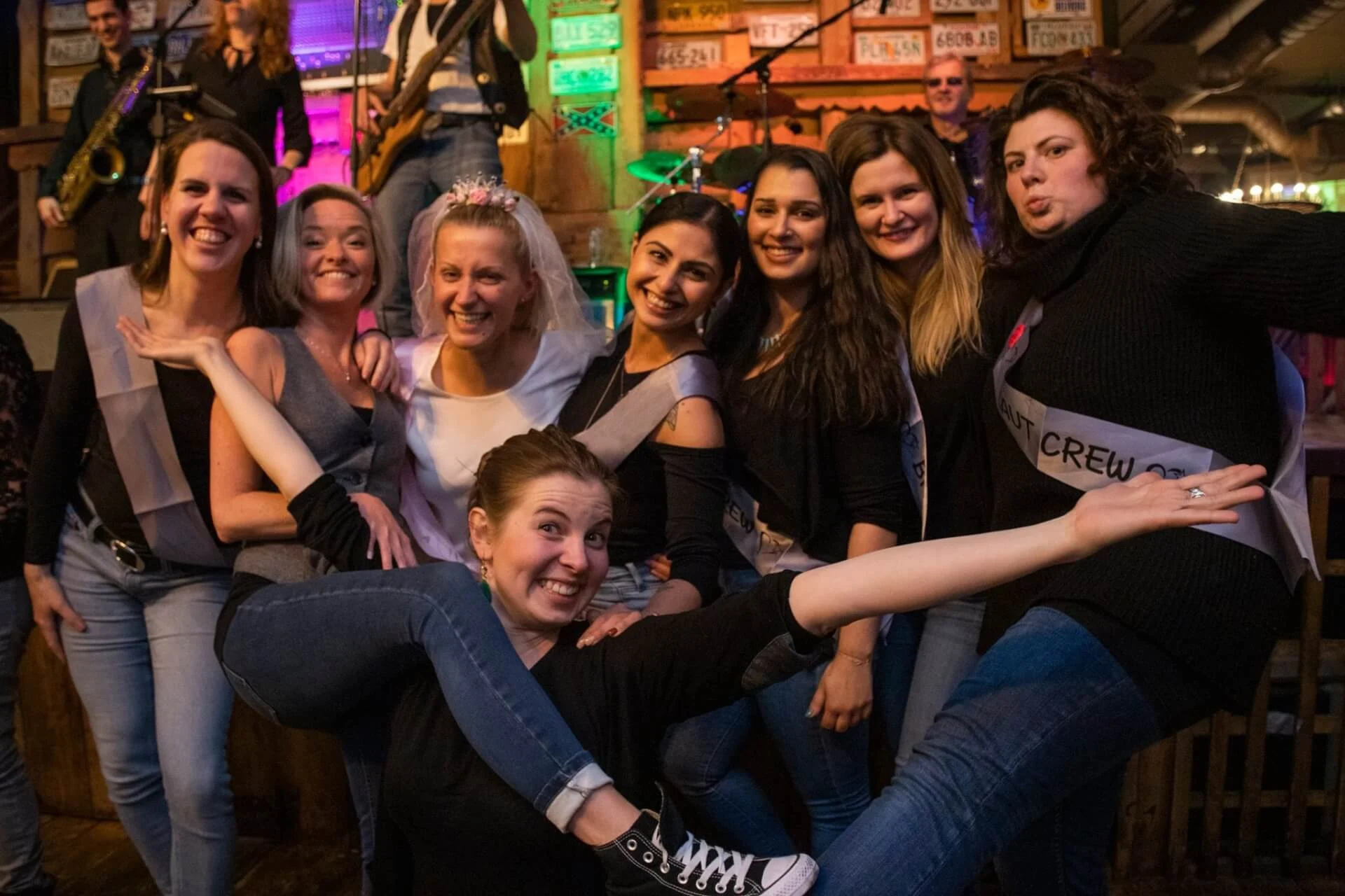 Eine Gruppe lächelnder Frauen posiert zusammen in einem Innenraum auf einer lebhaften Party, wobei eine Frau ein Diadem trägt. Eine Frau streckt spielerisch ihre Arme und Beine nach vorne, während im Hintergrund eine Band unter bunten Lichtern auftritt.