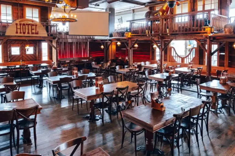 Geräumiges, rustikales Restaurant mit Holztischen und -stühlen, warmer Beleuchtung, rot getönten Stühlen, einem Balkon und einer Einrichtung im Western-Stil, einschließlich eines großen HOTEL-Schildes an der Wand.