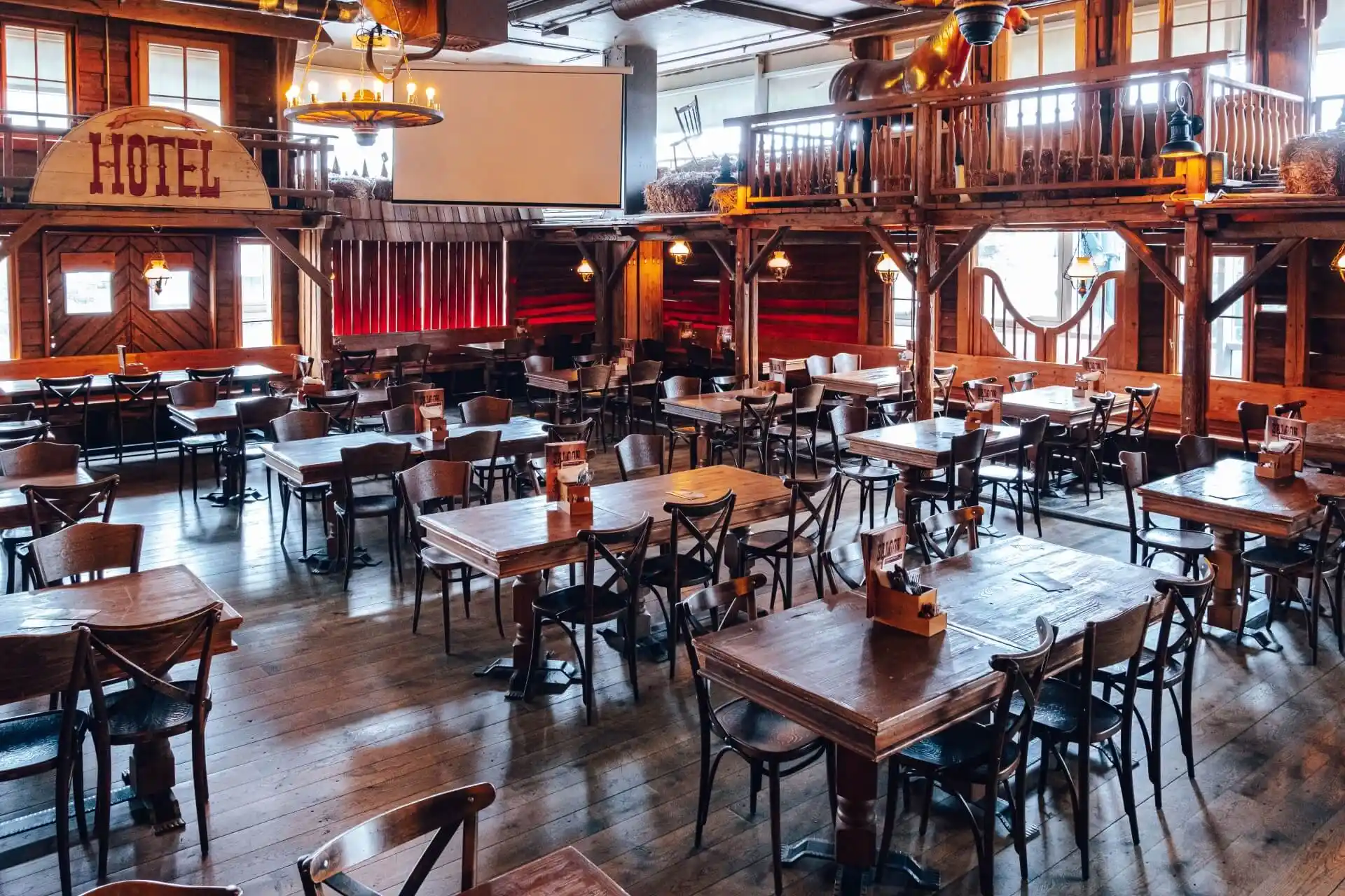 Geräumiges, rustikales Restaurant mit Holztischen und -stühlen, warmer Beleuchtung, rot getönten Stühlen, einem Balkon und einer Einrichtung im Western-Stil, einschließlich eines großen HOTEL-Schildes an der Wand.