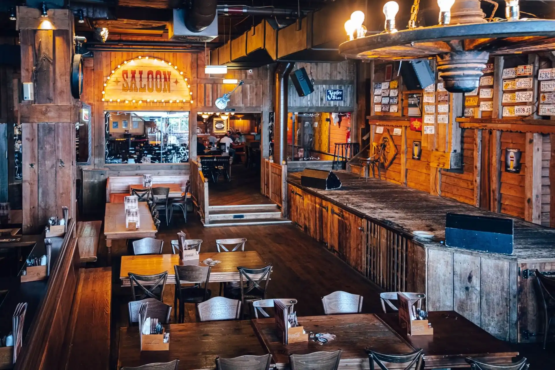Ein rustikaler, hölzerner Saloon mit einer Bühne, Holztischen und -stühlen, einem gut beleuchteten "Saloon"-Schild und warmer Umgebungsbeleuchtung erinnert an die Atmosphäre des Alten Westens.