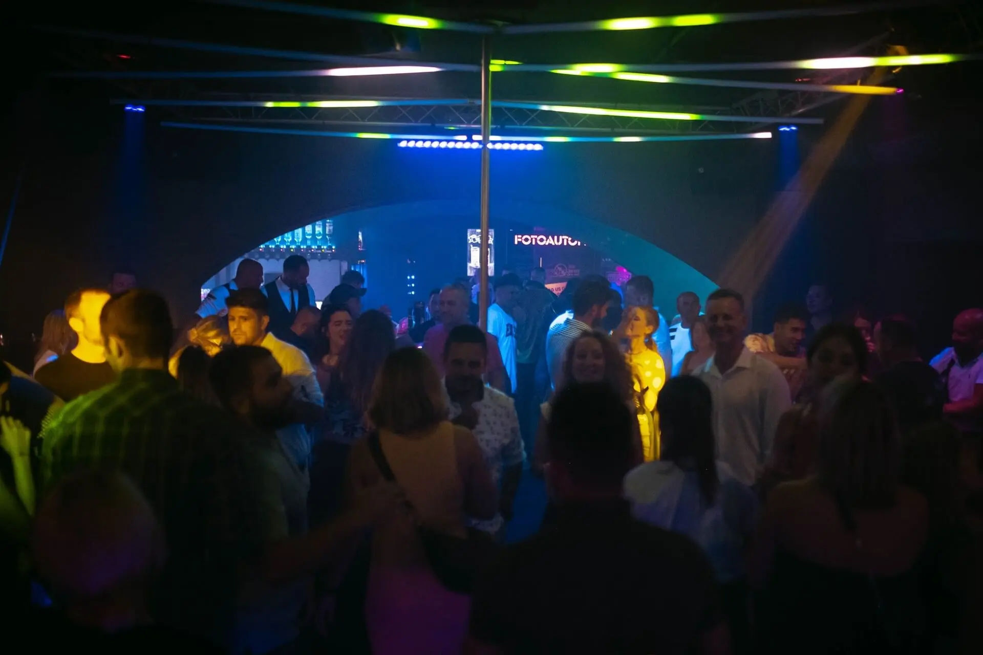 Eine Gruppe von Menschen tanzt und unterhält sich unter bunten Lichtern in einem überfüllten Nachtclub, mit einer Stange in der Mitte und einem neonbeleuchteten Barbereich im Hintergrund.