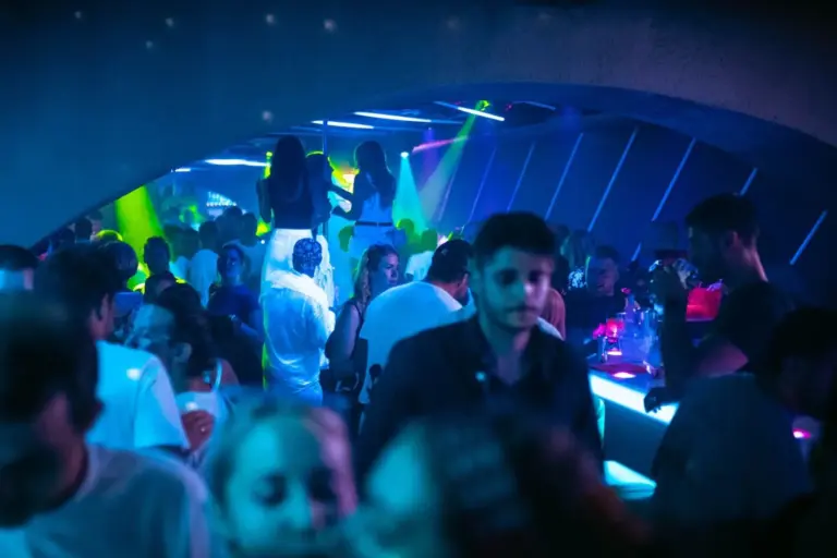 Belebte Nachtclubszene mit tanzenden und sprechenden Menschen unter blauen und violetten Neonröhren. Einige stehen an der Bar, andere tanzen auf erhöhten Plattformen und schaffen eine lebhafte, energiegeladene Atmosphäre.