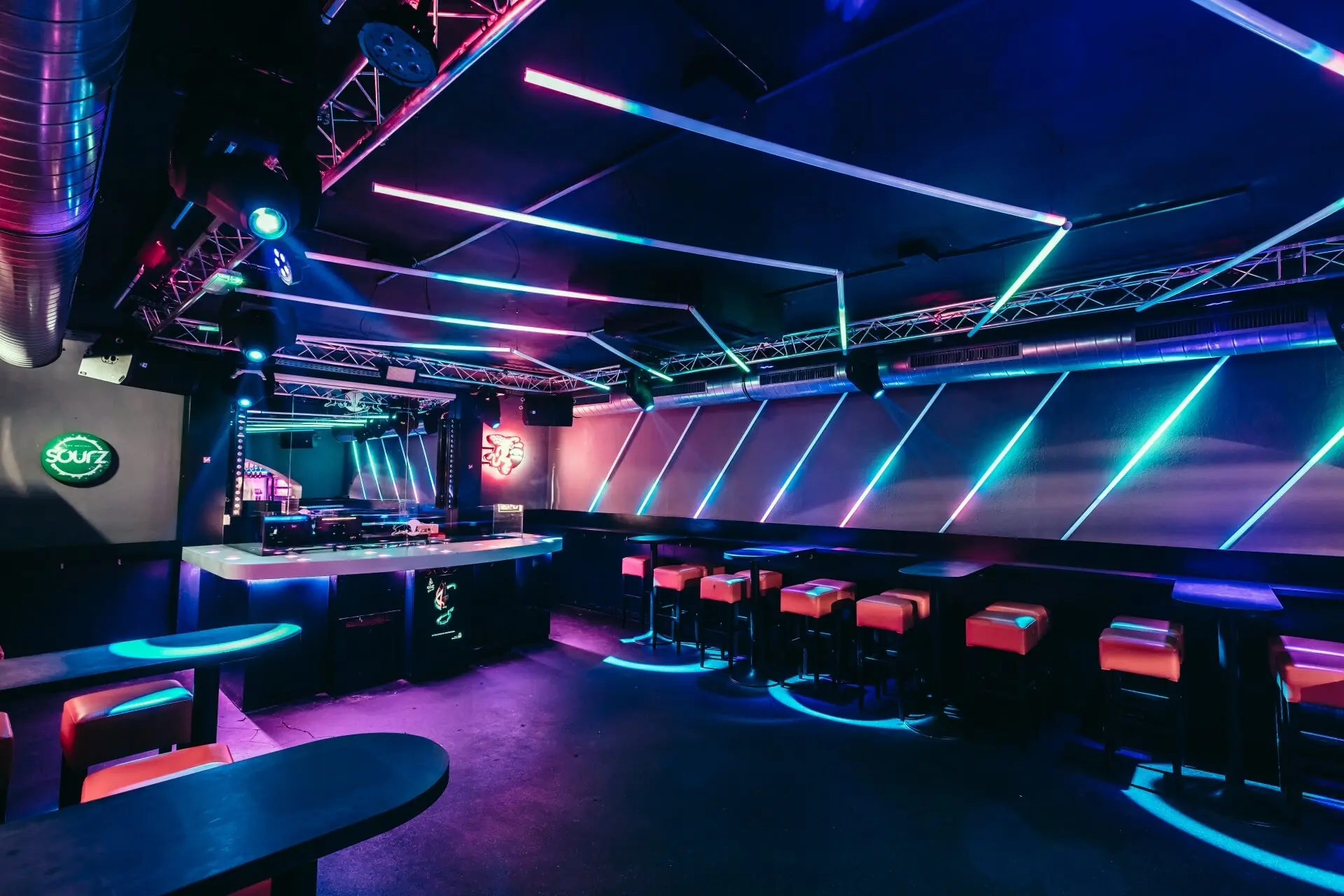 Ein moderner Nachtclub mit neonblauer und rosafarbener Beleuchtung, schwarzen Tischen, rot gepolsterten Barhockern und einer DJ-Kabine im Hintergrund, die eine lebendige und futuristische Atmosphäre schaffen.