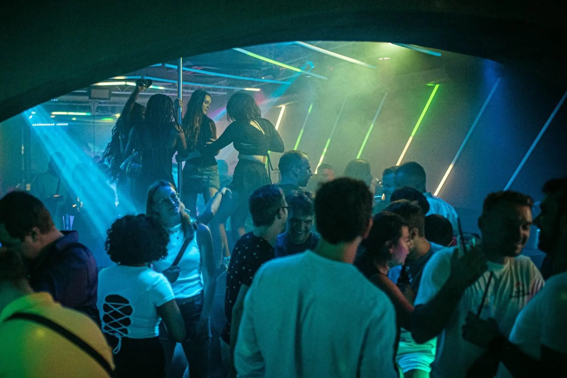 Eine belebte Nachtclubszene mit Menschen, die tanzen, sich unterhalten und sich unter bunten Neonlichtern und einer rauchigen Atmosphäre amüsieren. Einige Personen stehen auf einer Plattform über der Menge.