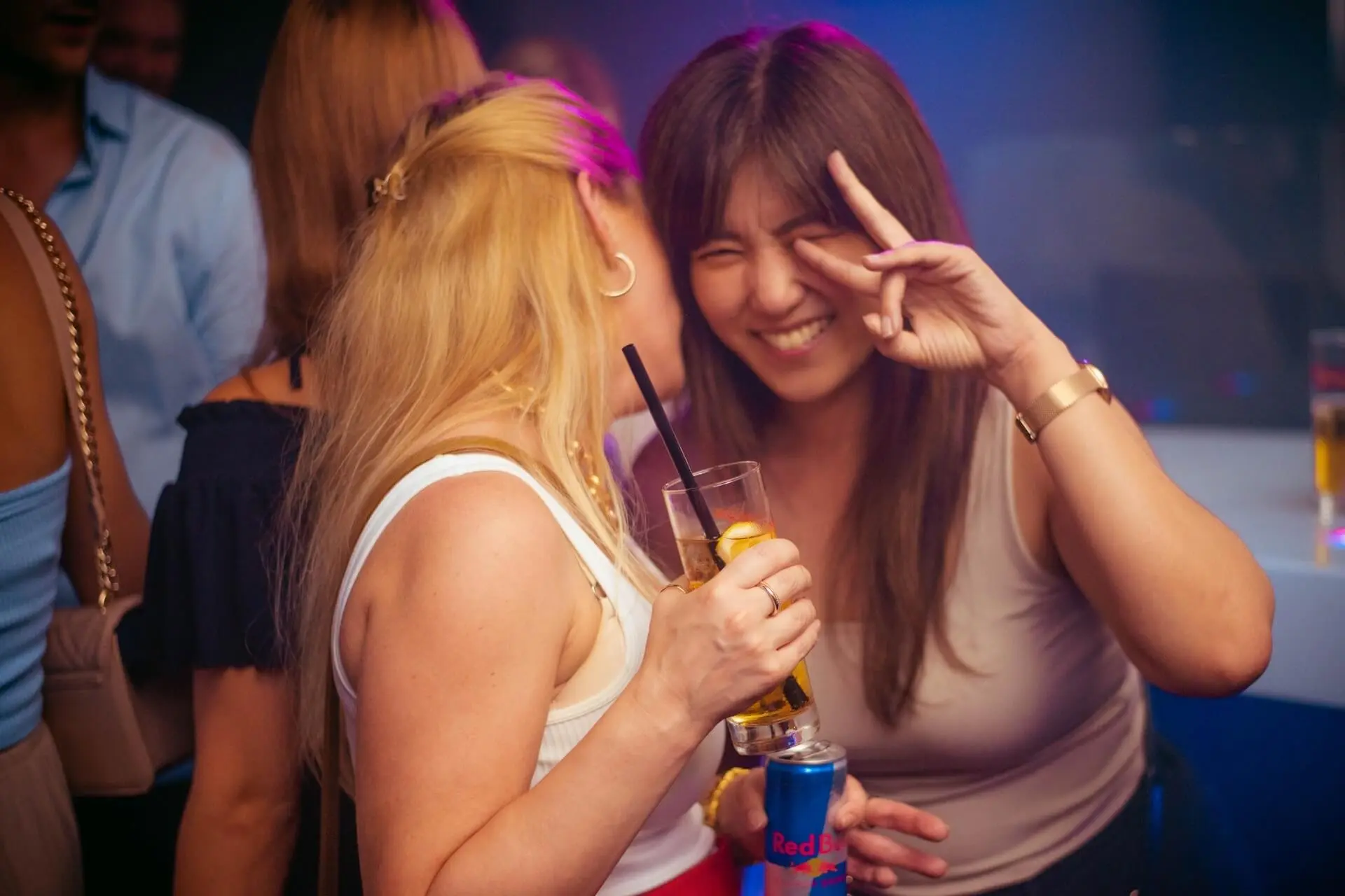 Zwei Frauen auf einer Party; eine flüstert, während sie ein Getränk in der Hand hält, die andere lächelt und macht ein Friedenszeichen mit ihren Fingern. Im Hintergrund sind Menschen und bunte Lichter zu sehen.
