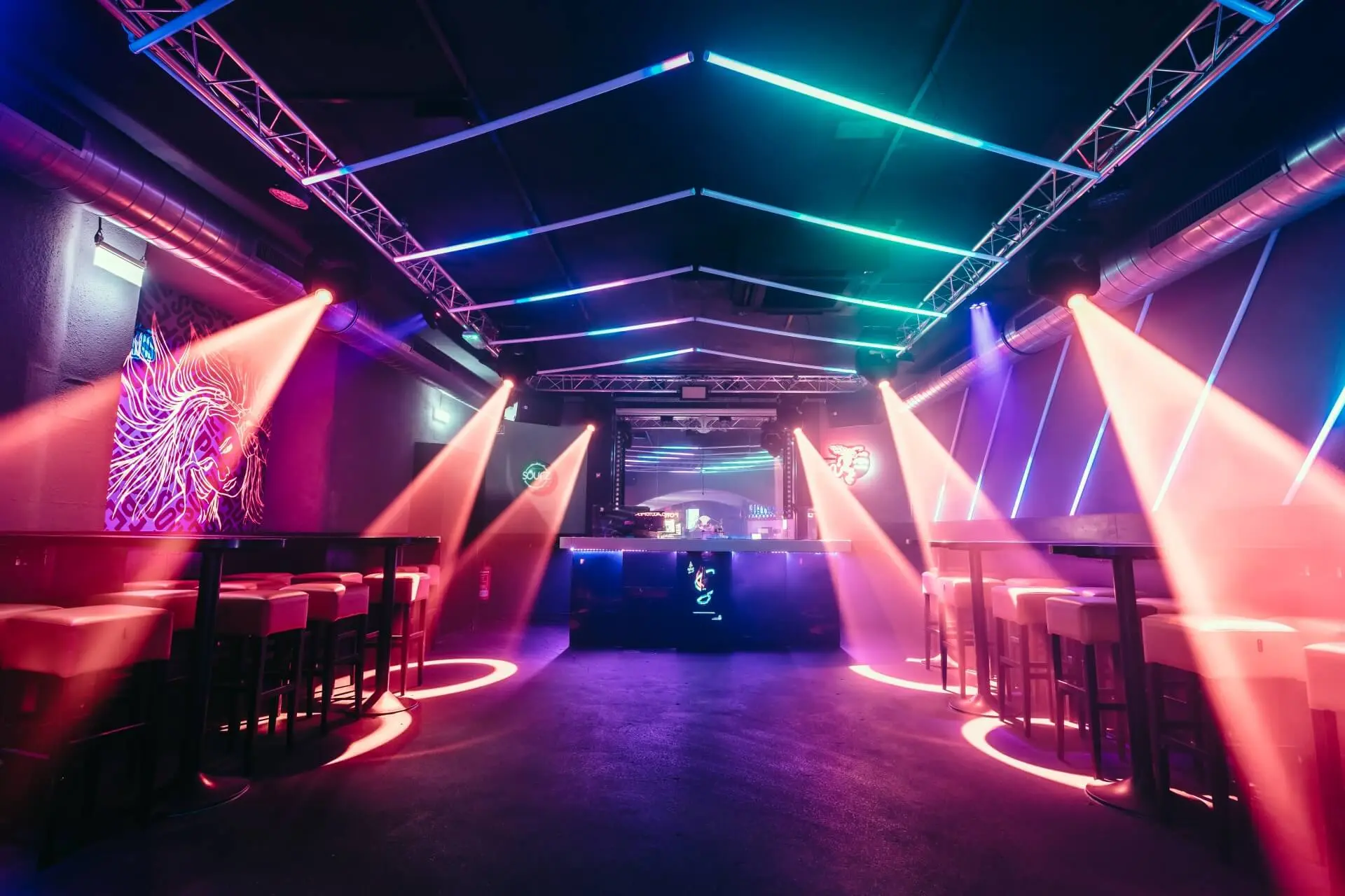 Ein moderner Nachtclub mit bunten Neonlichtern an der Decke, rosafarbenen Scheinwerfern, die auf leere Tische und Barhocker scheinen, und einem DJ-Pult im hinteren Teil. Die Atmosphäre ist lebendig und futuristisch.