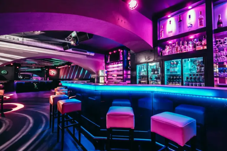 Ein moderner Nachtclub mit neonlila und blauer Beleuchtung, einem leuchtenden Bartresen, gepolsterten Barhockern, mit Flaschen bestückten Regalen und schlichtem Dekor schafft eine lebendige, futuristische Atmosphäre.