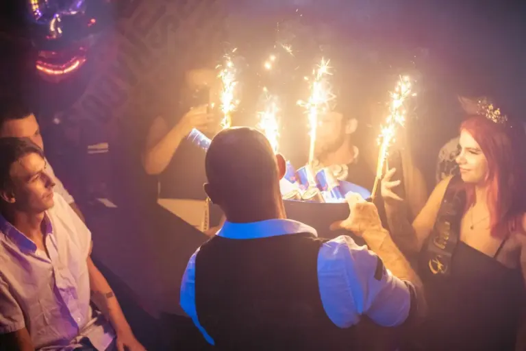 Eine Gruppe von Menschen auf einer schwach beleuchteten Party beobachtet, wie ein Mann in einer Weste ein Tablett mit Flaschen und Wunderkerzen präsentiert, die in der verrauchten Atmosphäre helle, festliche Funken erzeugen.