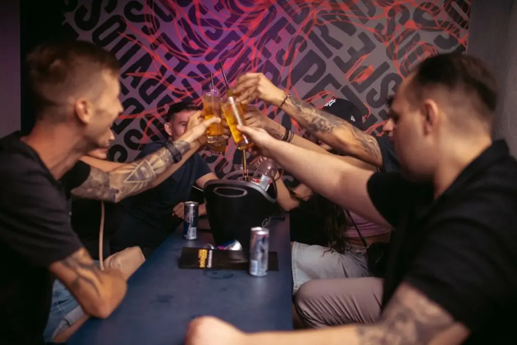 Eine Gruppe von Menschen mit Tattoos sitzt um einen Tisch herum und stößt mit ihren Getränken auf einen lebhaften Junggesellenabschied in einer modernen Bar mit bunter Wandkunst im Hintergrund an.