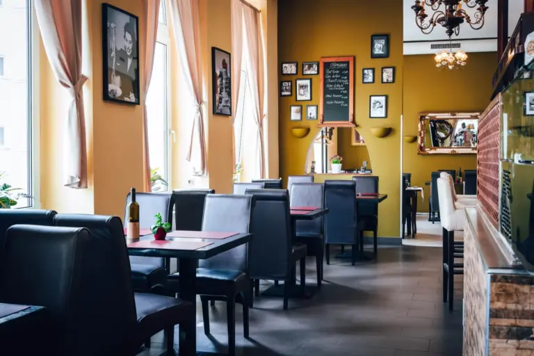 Ein gemütliches Restaurant mit schwarzen Tischen und Stühlen, großen Fenstern mit durchsichtigen Vorhängen, gerahmten Fotos an gelben Wänden und einer Speisekarte auf Kreidetafeln. Der Raum ist sanft beleuchtet und einladend.