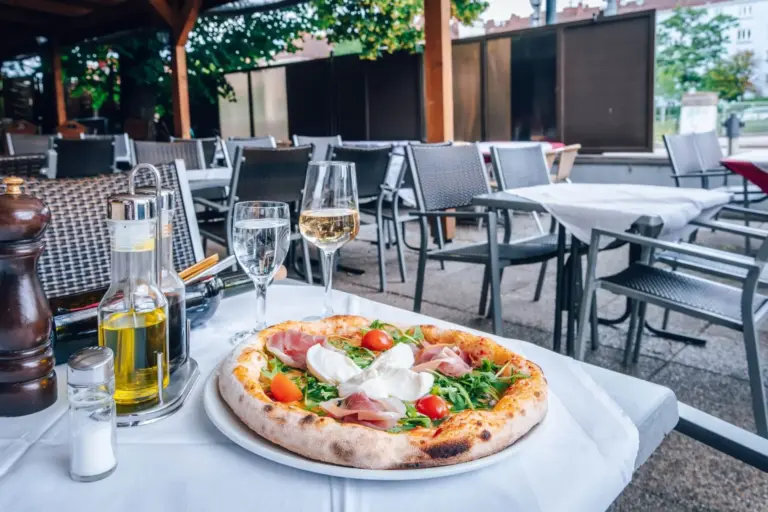 Eine frische Pizza mit Rucola, Prosciutto, Kirschtomaten und Burrata liegt auf einem Tisch mit Wein, Wasser und Gewürzen in einem Außenrestaurant mit leeren Stühlen und Tischen im Hintergrund.