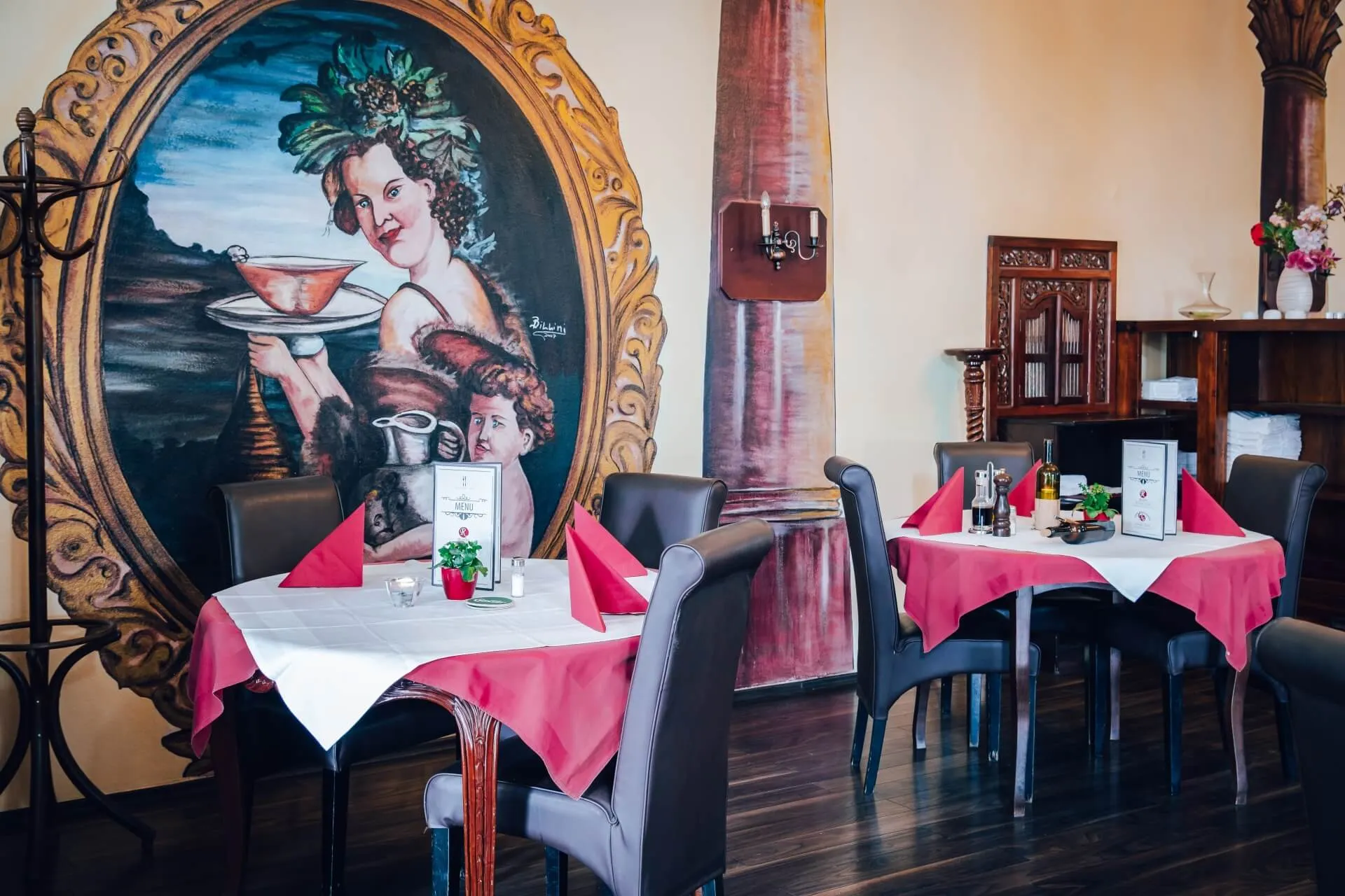 Ein gemütliches Restaurant mit Tischen, die mit roten und weißen Tischdecken, Speisekarten und Gewürzen gedeckt sind. Ein großes Wandgemälde mit einer Frau und einem Kind, die Getränke in der Hand halten, schmückt die Wand im Hintergrund.
