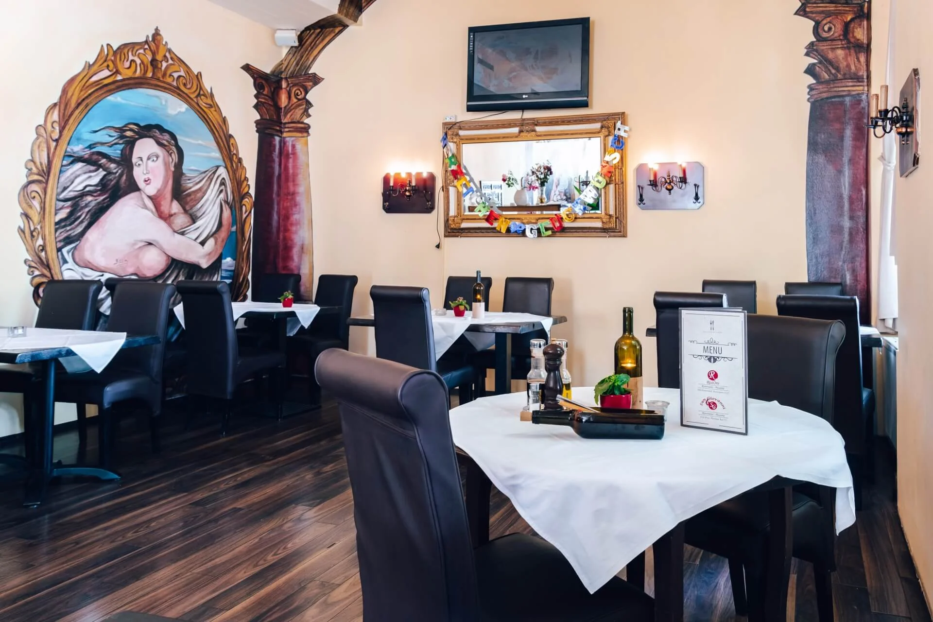 Ein gemütlicher Restaurantbereich mit dunklen Stühlen, weißen Tischdecken und ordentlich gedeckten Tischen. An den Wänden hängen ein großes Wandgemälde einer Frau und verzierte Spiegel, die eine warme und einladende Atmosphäre schaffen.