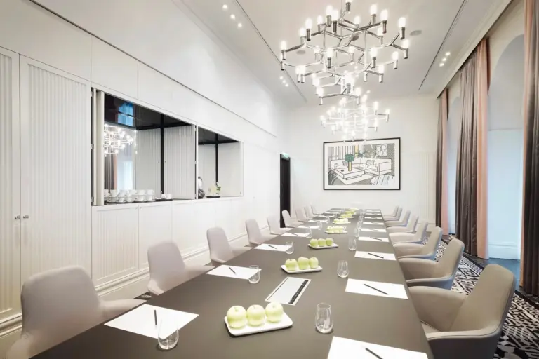 Ein moderner Konferenzraum mit einem langen schwarzen Tisch, der mit Notizblöcken, Stiften, grünen Äpfeln und Gläsern gedeckt ist. Beigefarbene Stühle umgeben den Tisch. Von der Decke hängen Kronleuchter, und die weißen Wände sind mit Kunstwerken geschmückt.