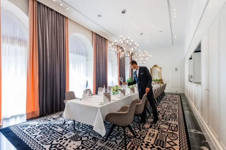 Ein Mann im Anzug deckt einen langen, elegant gedeckten Esstisch in einem hellen, modernen Raum mit großen Fenstern, Kronleuchtern und gemustertem Teppich.