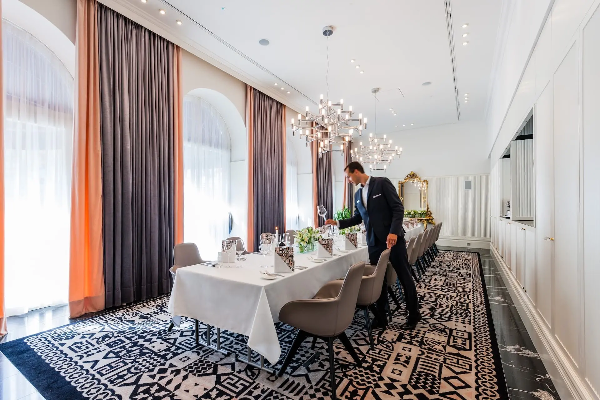 Ein Mann im Anzug deckt einen langen, elegant gedeckten Esstisch in einem hellen, modernen Raum mit großen Fenstern, Kronleuchtern und gemustertem Teppich.