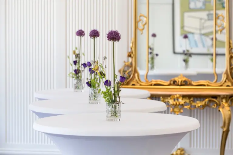 Drei runde Tische mit weißen Tischtüchern sind jeweils mit einer kleinen Glasvase mit violetten Blumen geschmückt. Dahinter befinden sich ein verschnörkelter goldgerahmter Spiegel und weiß getäfelte Wände mit einem gerahmten Kunstwerk, das sich im Spiegel spiegelt.