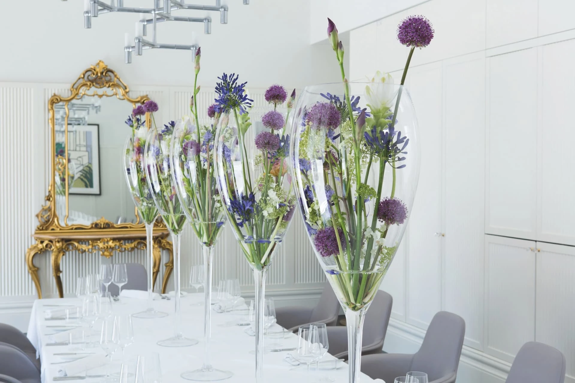 Ein langer Esstisch ist mit großen, dekorativen Glasvasen gedeckt, die mit lila und weißen Blumen gefüllt sind. Elegante Stühle und Gedecke umgeben den Tisch. An der Wand im Hintergrund hängt ein goldener, verzierter Spiegel.