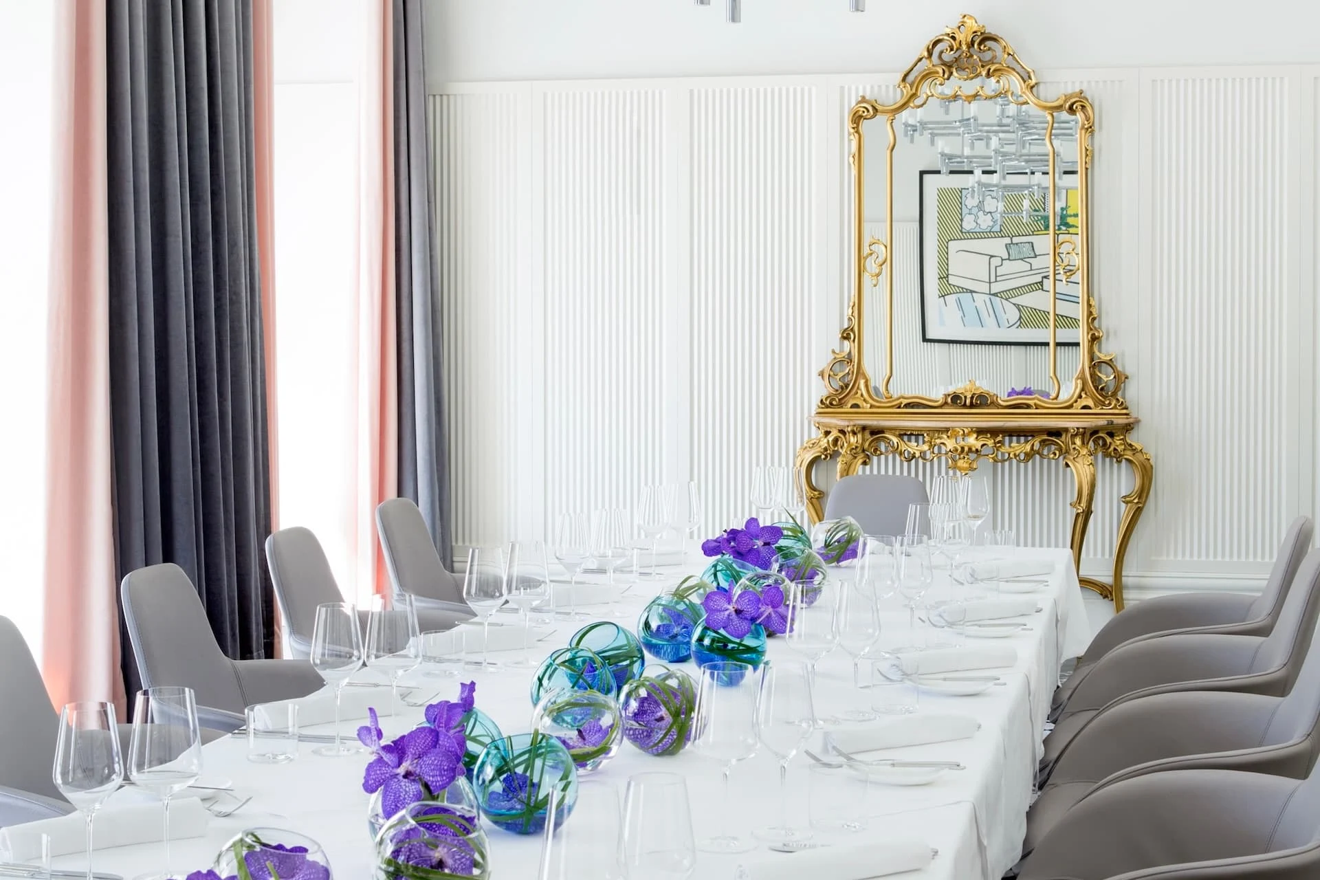 Ein langer Esstisch mit weißem Tischtuch ist für eine Mahlzeit gedeckt, mit klaren Gläsern und Tafelaufsätzen mit lila Blumen. Graue Stühle säumen den Tisch, und ein großer, verzierter goldener Spiegel hängt an der weißen Wand dahinter.
