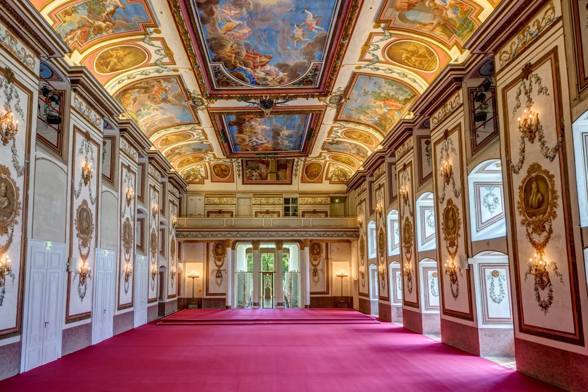 Großer Saal mit leuchtend rotem Teppich, kunstvollen Goldverzierungen, großen Fenstern und einer aufwendig bemalten Decke mit klassischer Kunst und verschlungenen Mustern, beleuchtet von natürlichem Licht durch offene Türen und Fenster.