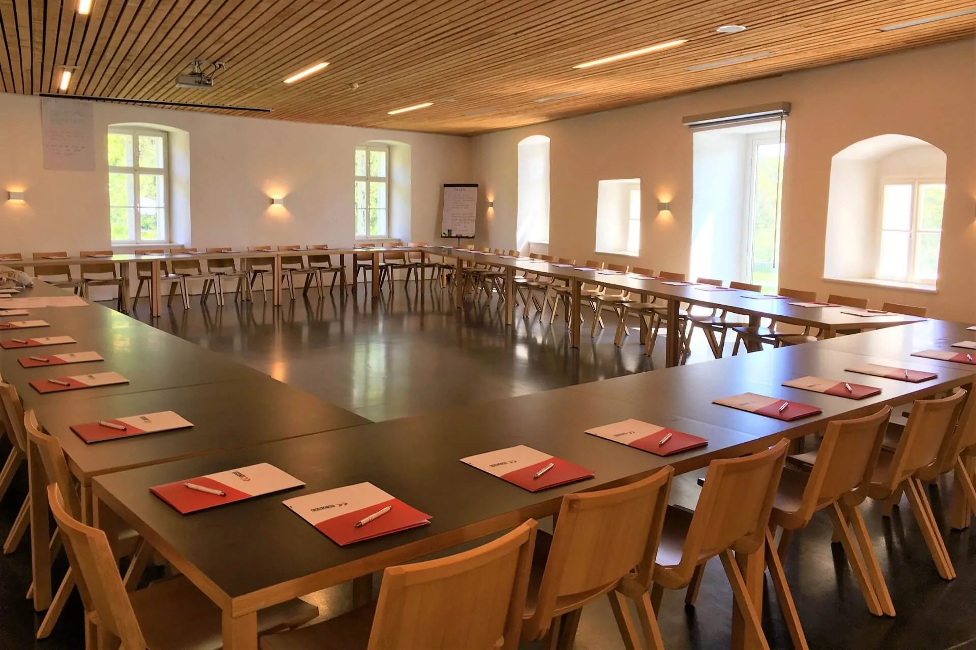 Ein geräumiger, gut beleuchteter Konferenzraum mit Holzstühlen, die in einer großen U-Form um Tische angeordnet sind. Jeder Platz ist mit einem roten Notizbuch und einem Stift ausgestattet. Große Fenster säumen die Wände, und in der Ecke steht ein Flipchart.