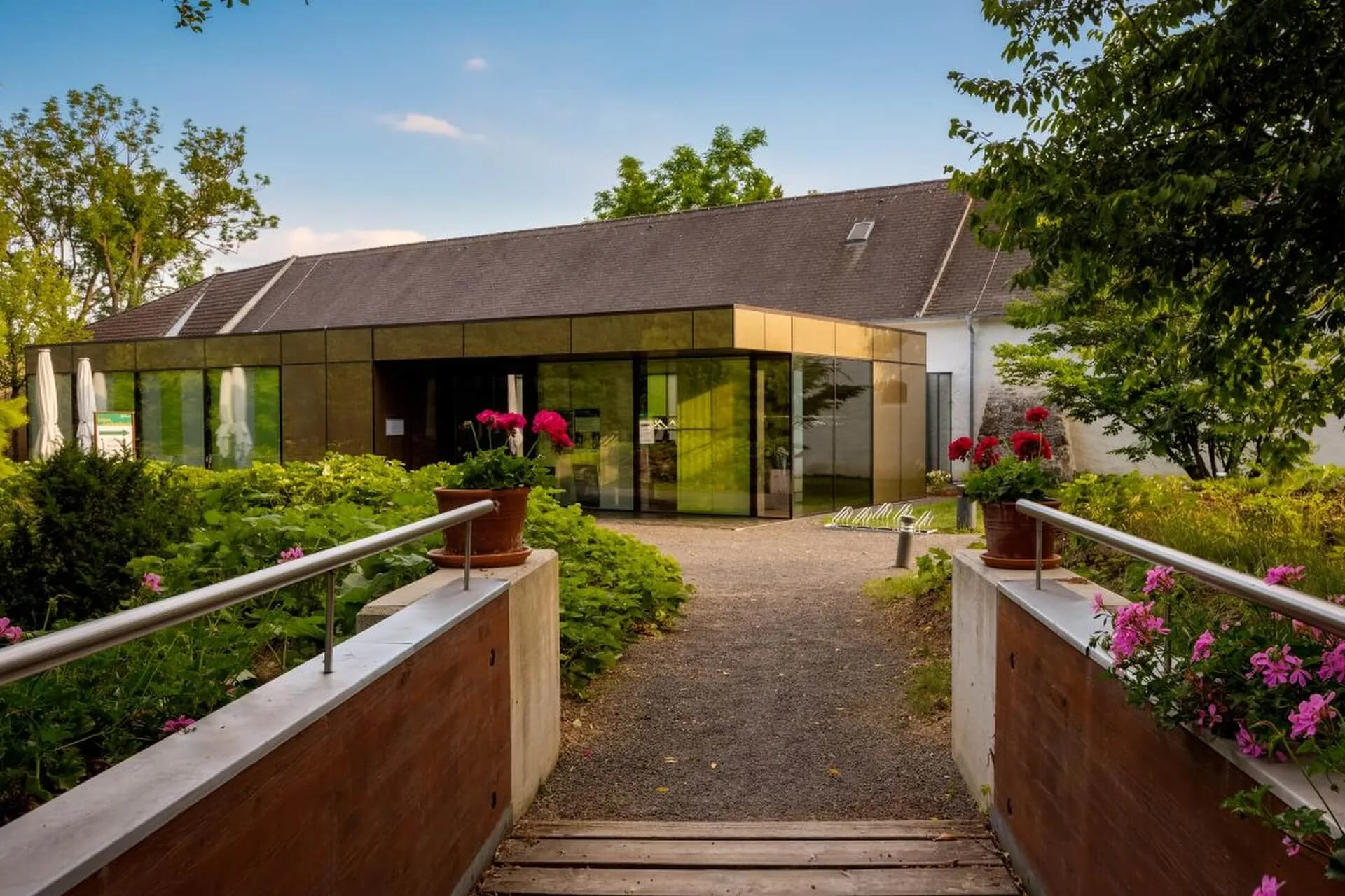 Ein modernes gläsernes Gebäude mit einem schrägen Dach ist von grünen Sträuchern und Bäumen umgeben. Eine kleine Holzbrücke mit Blumen in Töpfen führt unter blauem Himmel zum Eingangsweg.