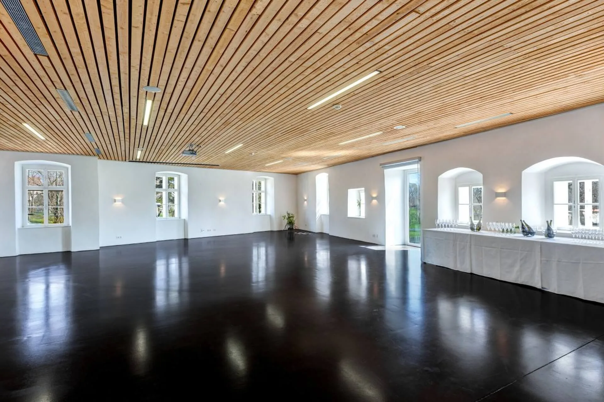 Geräumiger, moderner Veranstaltungsraum mit Holzlattendecke, weißen Wänden, schwarzem, glänzendem Boden, großen Fenstern und einem langen Tisch mit Gläsern und Flaschen in der Nähe der Fenster.