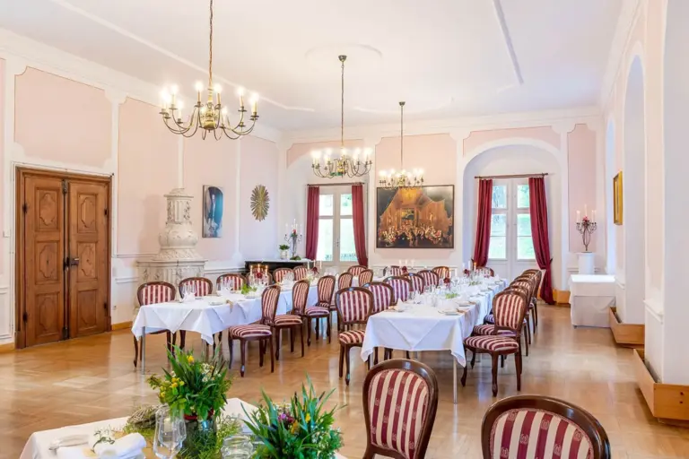 Eleganter Speisesaal mit Kronleuchtern, Tischen mit weißen Tischtüchern und Blumenschmuck, gestreiften Stühlen, großen Fenstern mit roten Vorhängen, Holzfußboden und klassischen Kunstwerken an rosa Wänden.