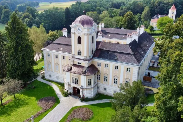 Luftaufnahme eines großen, verschnörkelten, blassgelben Herrenhauses mit einer zentralen Kuppel, umgeben von üppigen grünen Rasenflächen, Bäumen und einem gewundenen Weg in einer ländlichen Landschaft.